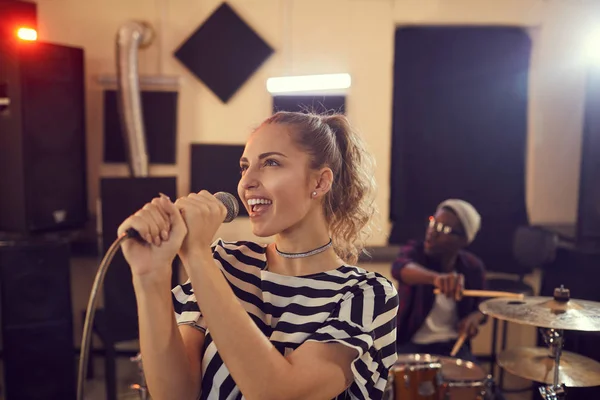 Portrait of smiling young woman singing to microphone during rehearsal or concert with music band in background, copy space