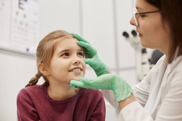 Portrait of female optometrist checking eyes of smiling little girl in modern ophthalmology clinic, copy space