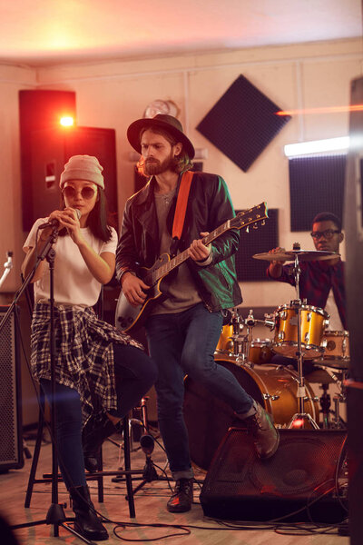 Full length portrait of contemporary band playing music during rehearsal or concert in dimly lit studio, focus on handsome man playing guitar in foreground, copy space