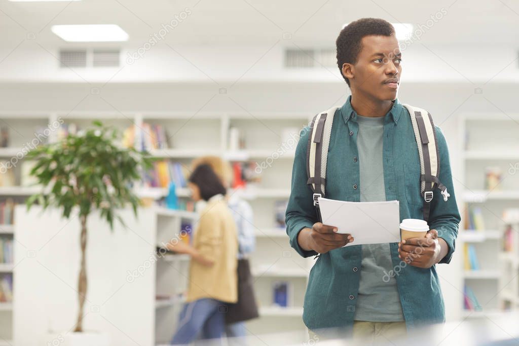 Waist up portrait of African-American student holding coffee cup while standing in college library, copy space