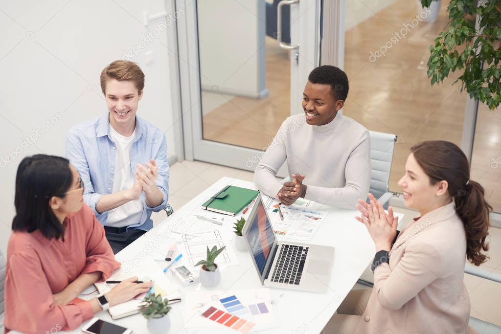 High angle view at multi-ethnic group of business people clapping to Asian colleague while collaborating on design project during meeting in office, copy space