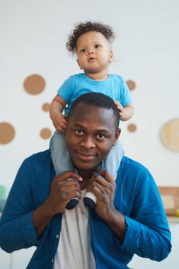 Omuzlarında oğlunu taşıyan ve kameraya bakıp gülümseyen mutlu Afro-Amerikan bir adamın dikey portresi.