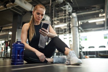 Modern genç bir kadının spor salonunda spor salonunda otururken akıllı telefon kullanmasının ve antremana ara vermesinin tam boy portresi.