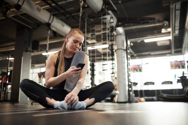 Jimnastik salonunda Lotus pozisyonunda otururken akıllı telefon mesajlarını kontrol eden sarışın genç bir kadının tam boy portresi.