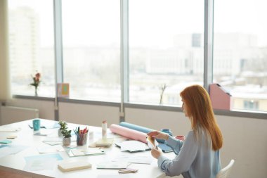 İş yerinde pencere kenarında otururken elinde renk örnekleri tutan kızıl saçlı kadın tasarımcının geniş açı portresi, boşluğu kopyala