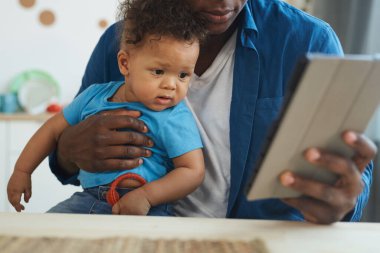 Ev içinde dijital tablet kullanırken bebeği tutan Afro-Amerikalı bir adamın kesilmiş portresi, boşluğu kopyala