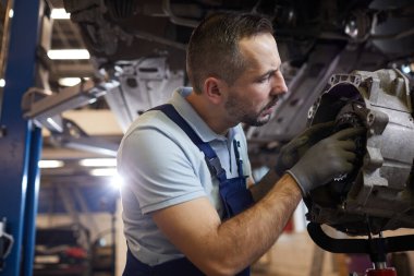 Otomobil atölyesinde vites tamir eden sakallı araba tamircisinin yan görünüm portresi.