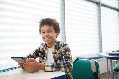 Okul sınıfında masa başında oturmuş kameraya bakarken elinde akıllı telefon tutan neşeli Afro-Amerikan çocuk portresi.