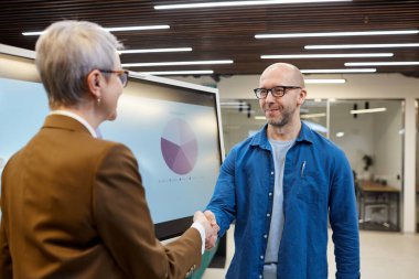 Ofiste sunum panosuna karşı dururken iş kadınıyla tokalaşan kel bir adamın belden yukarı portresi. Boşluğu kopyala.