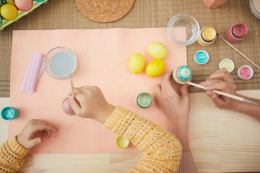 Üst manzara, anne ve kızı Paskalya yumurtalarını boyuyor. Pastel renkler mutfak masasında, iç kısımda oturuyor, boşluğu kopyalıyor.