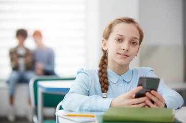 Okul sınıfında masada oturmuş kameraya bakarken akıllı telefonu tutan tatlı sarışın kızın portresi.