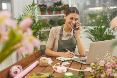 Çiçekçide telefonla konuşurken kameraya gülümseyen kadın küçük işletme sahibinin portresi.