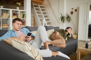 Evdeki kanepede televizyon izleyen, ön planda akıllı telefon kullanan bir adama odaklanan sıkılmış genç bir çiftin tam boy portresi.