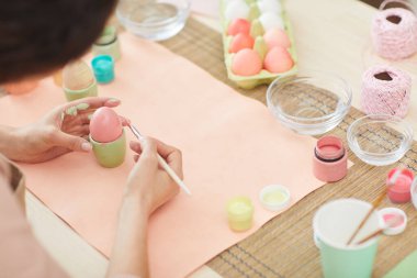 Yüksek açılı, tanınmayan bir kadının paskalya yumurtalarını pastel renklerle boyarken mutfak veya sanat atölyesinde otururken görüntüsü.