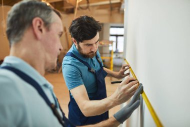 Evi yenilerken duvarları ölçen iki inşaat işçisinin belden yukarı portresi.