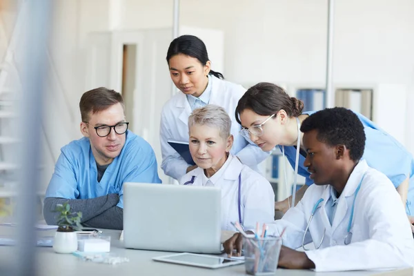 医療室内の机に座りながらノートパソコンを使っている多民族の医師たち — ストック写真