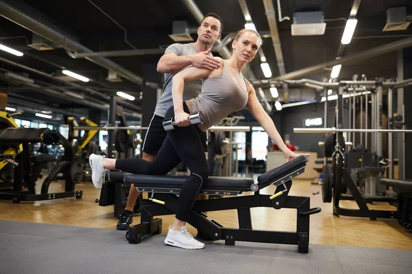 Full length portrait of muscular coach correcting position of young woman doing exercises with dumbbells during strength workout in modern gym, copy space