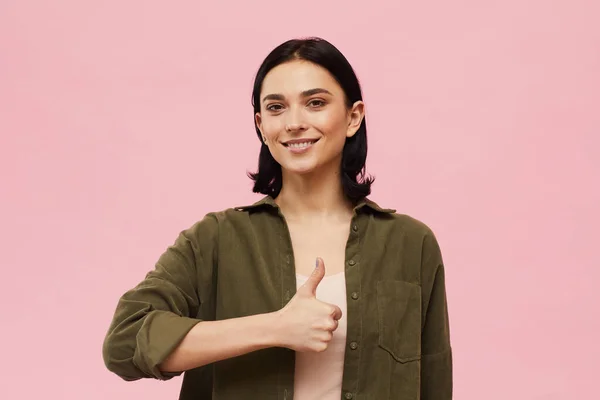 Waist up portrait of modern young woman showing thumbs up and looking at camera while standing against pink background in studio, copy space