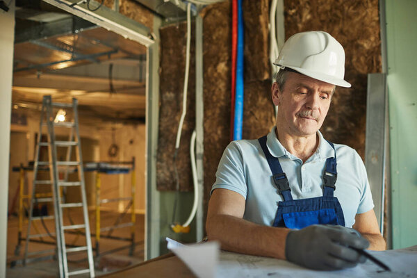 Waist up portrait of senior construction worker wearing hardhat and looking at floor plans while renovating house, copy space