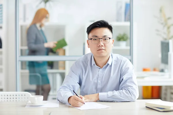 Portrait of young Asian businessman wearing glasses and looking at camera while posing at workplace in office cubicle, copy space