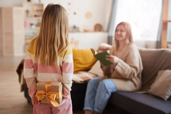 Back view portrait of cute girl hiding gift for mom while surprising her on Mothers day or Birthday in cozy home interior, copy space