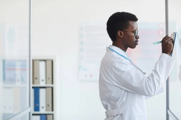 Side view portrait of young African-American doctor writing on clipboard while standing by glass wall in med clinic, copy space