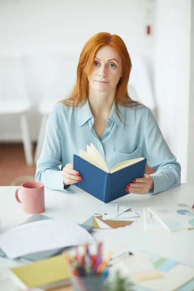 Portrait of freckled young woman looking at camera while holding planner at designers workplace