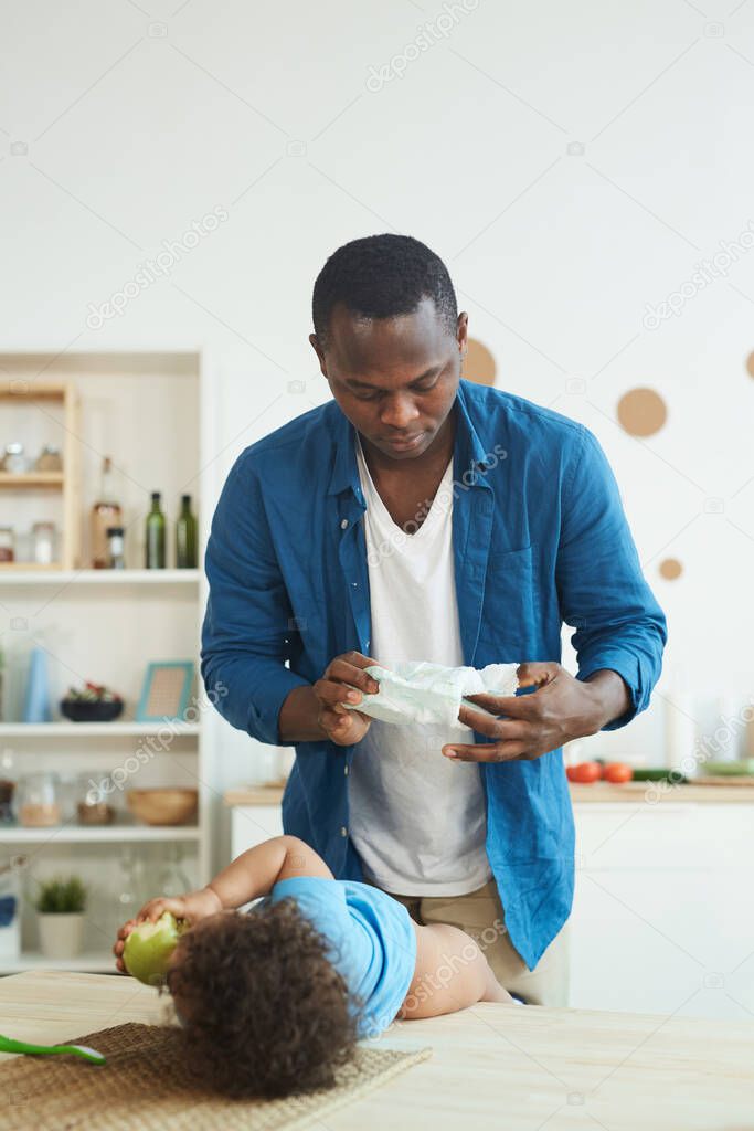 Vertical portrait of adult African-American man changing diaper while caring for toddler at home, copy space