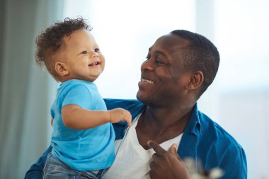 Mutlu Afro-Amerikan baba portresi. Gülen küçük çocukla oynuyor. Mutfak masasında oturan beyaz çocuk.