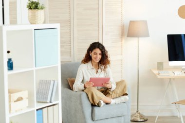 Genç bir kadın koltuğunda rahatça oturup modern tablet bilgisayar ve internet kullanarak işini yapıyor.
