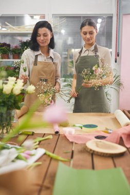 Çiçekçide çalışırken buketleri ve çiçekli kompozisyonları düzenleyen iki yaratıcı genç kadının dikey portresi.