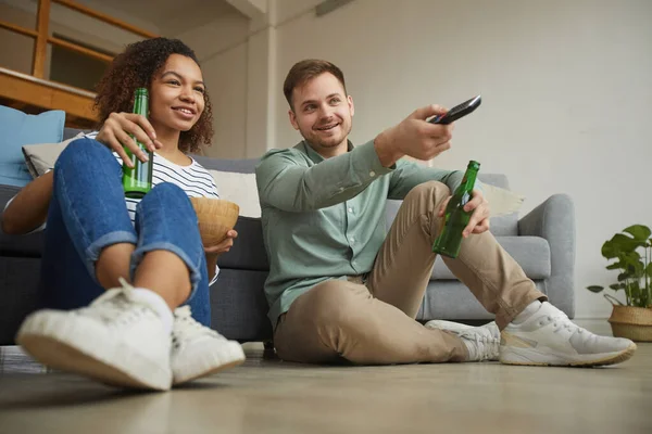 一对年轻的男女坐在舒适的公寓地板上看电视和喝啤酒的全长肖像画 复制空间 — 图库照片