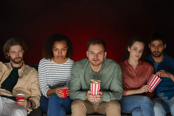 Group of friends watching movies at home while eating snacks and popcorn sitting on big sofa in dark room