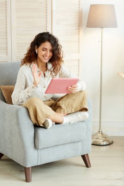 Evdeki koltukta rahatça oturan ve tablet bilgisayar ve kulaklık kullanarak video konferansına katılan genç bir kadın.