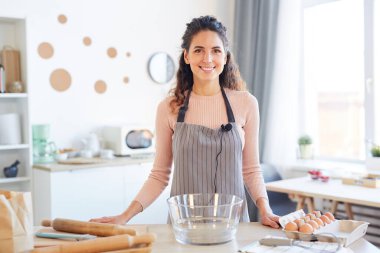Mutfak masasında durmuş gülümseyen kameraya bakarken önlük giymiş neşeli genç bir kadının yatay portresi.