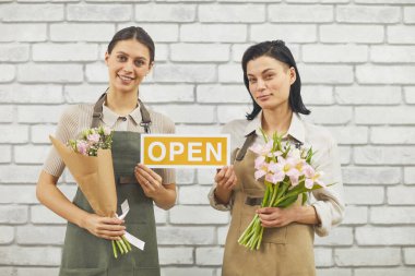 Açık tabela tutan ve elinde güzel bahar çiçekleriyle kameraya gülümseyen iki kadın çiçekçinin belden portresi.