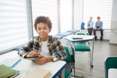 Sınıfta oturmuş kameraya bakarken elinde akıllı telefon tutan gülümseyen Afro-Amerikan çocuğun geniş açılı portresi.