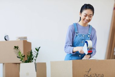 Asyalı genç bir kadının kasetle karton kutular paketleyip mutlu bir şekilde gülümsediği belden yukarı portresi. Yeni eve taşınmak için heyecanlı, kopyalama alanı.