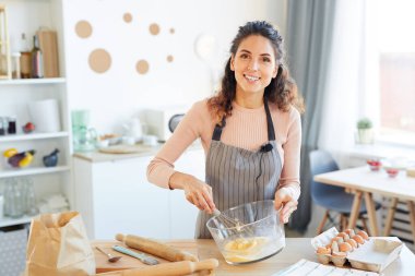Neşeli yemek blogcusu önlük takıyor modern mutfakta masada duruyor cam kasede yumurtaları kırbaçlıyor, kamera portresine bakıyor.