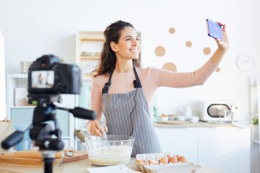 Önlük giymiş çekici genç bir kadın yemek videosu çekerken selfie çekiyor.