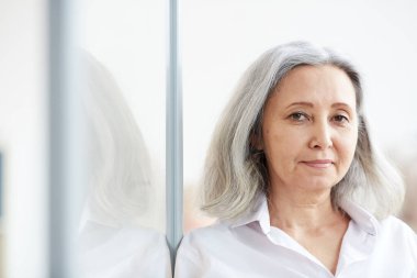 Ofiste cam duvara yaslanırken kameraya bakan kıdemli iş kadınının kafa ve omuz portresi.