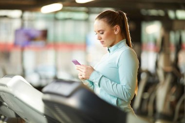 Spor salonundaki koşu bandında kardiyo çalışmasına hazırlanırken elinde akıllı telefon tutan genç bir kadının yan görünüş portresi.