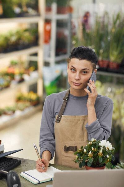 Waist up portrait of female small business owner speaking by smartphone and looking at camera while standing behind counter in flower shop