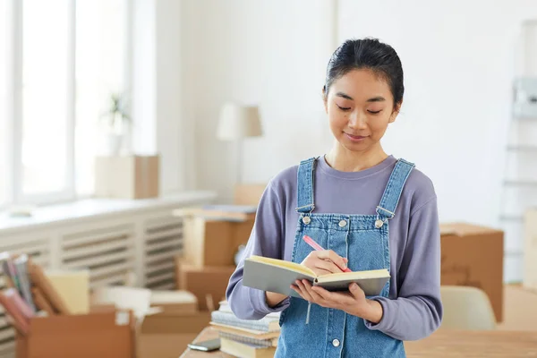 背景に段ボール箱付きの新しい家やアパートに立っている間 プランナーで書く若いアジアの女性のウエストアップ肖像画 コピースペース — ストック写真