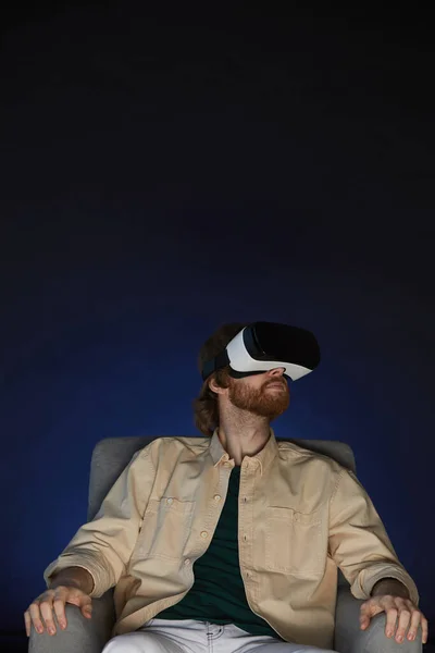 Vertical portrait of modern bearded man wearing virtual reality gear while enjoying immersive videogame or movie in dark, copy space