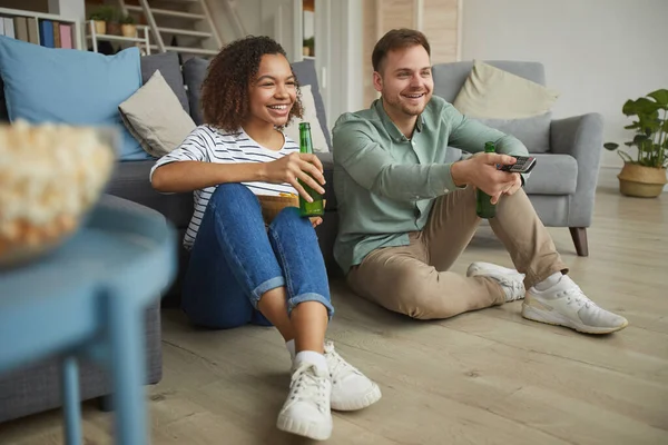 Modern melez bir çiftin tam boy portresi. Evde televizyon izleyip, yerde otururken bira içiyorlar.