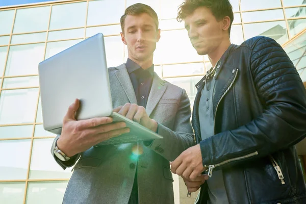 Low angle portrait of two handsome young men using laptop outdoors discussing work while standing outside of modern office building, lens flare — entrepreneurs, male - Stock Photo | #181813690