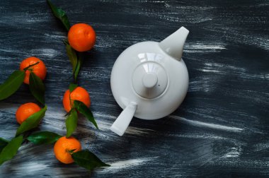 Beyaz seramik çaydanlık ve koyu ahşap arka plan üzerinde çuval bezi üzerinde yeşil yaprakları ile mandalina 