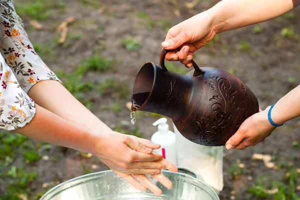 Mãos de uma mulher segurando um jarro autêntico de barro e derramado nas mãos de uma jovem, o conceito de relaxar ao ar livre — Fotografia de Stock