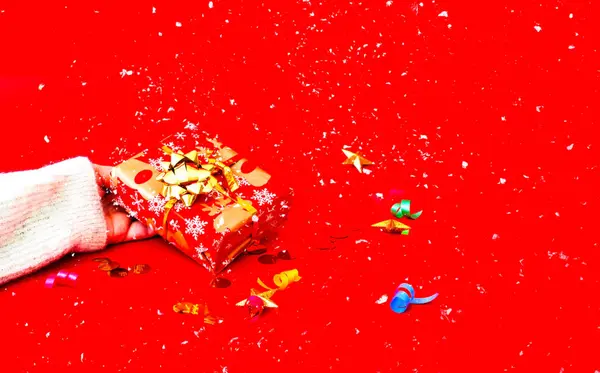 Mão feminina detém presente de Natal em embalagens festivas em fundo vermelho com confete. Espaço de cópia — Fotografia de Stock